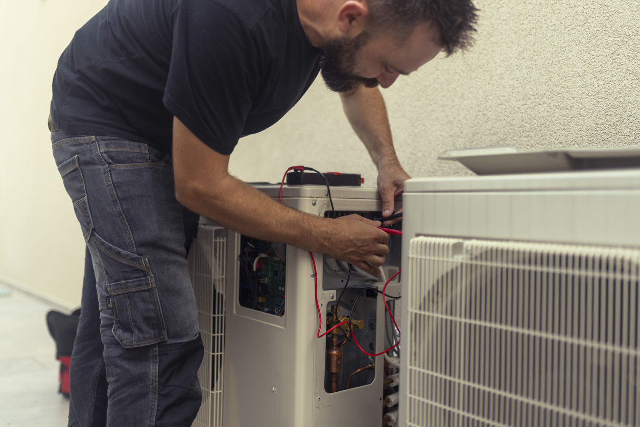 An HVAC technician kneels beside an outdoor air conditioner unit, using red and black multimeter probes to test its internal wiring and components as he installs it.
