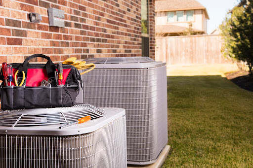 Bag of repairman's work tools, gloves on top of two air conditioner units outside a brick home in a residential neighborhood